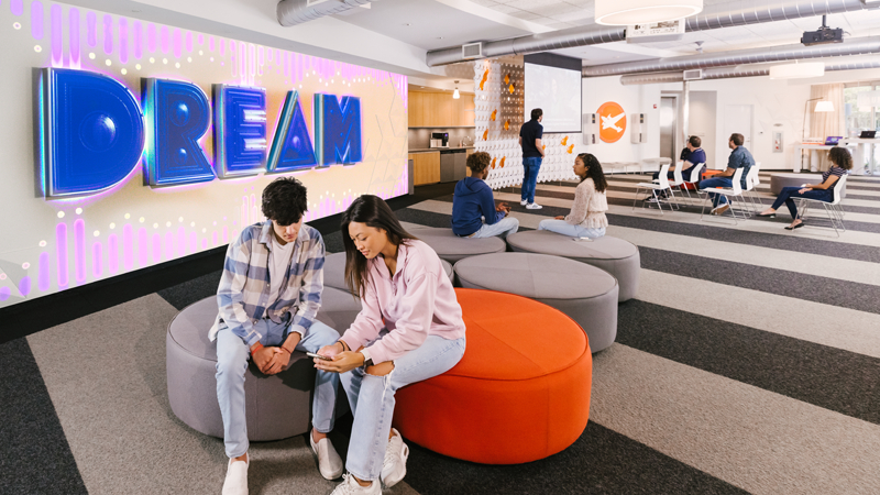 Students sit in Full Sail’s Welcome Center. There is a Dream sign on one of the walls and a staff member talking in front of a projection screen.