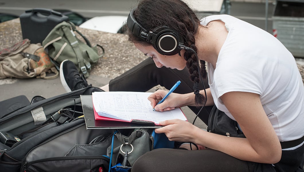 A student wearing headphones, a white shirt, and black pants sits on the ground next to a backpack. They are writing in a notebook.