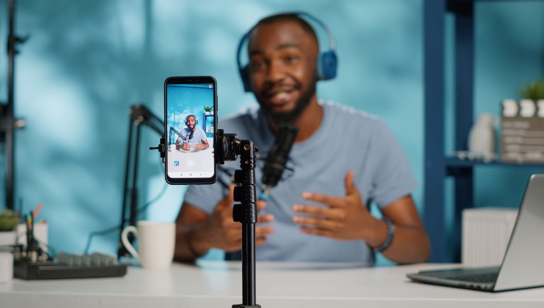 A person is seen out of focus while seated at a podcasting station, a smart phone is recording while mounted on a stand in the foreground.