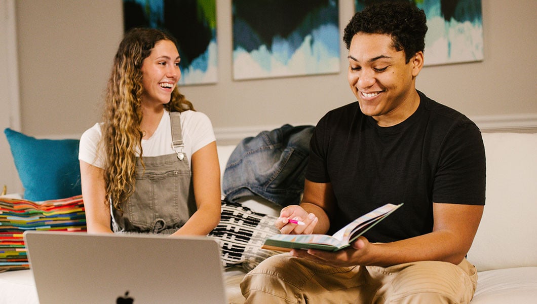 Two smiling students sit on a couch and work together in a living room. One student holds a book, and a laptop sits on the table in front of them.
